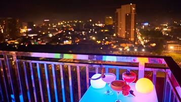 Un balcón con una mesa que tiene bebidas y una esfera brillante, con vistas a un paisaje urbano nocturno.