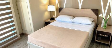 Una cama bien hecha con una manta color beige, una lámpara de noche y un jarrón con flores sobre una mesita de noche.