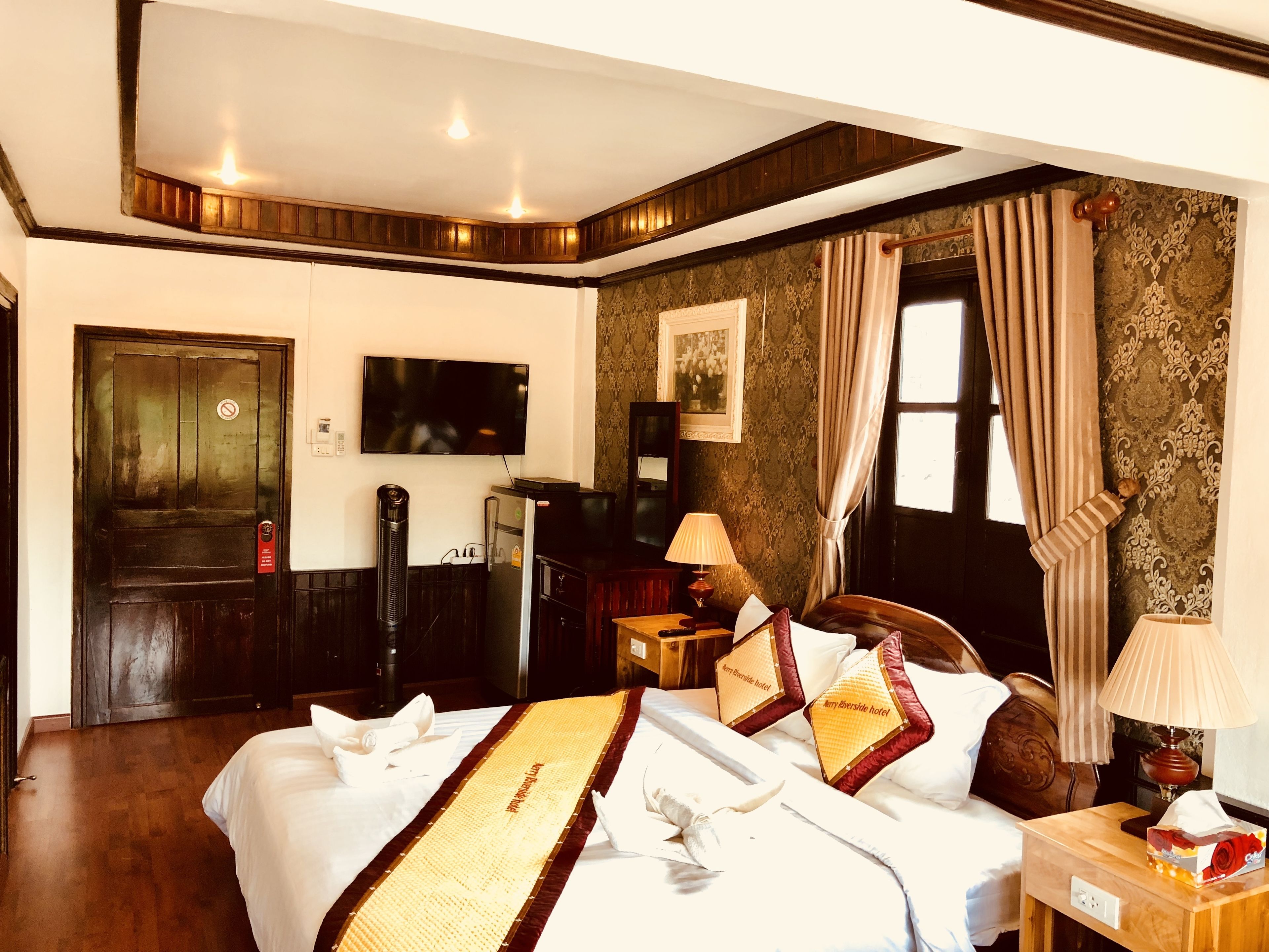 A hotel room with a bed, a television, a desk with a lamp, a window with curtains, and a door with a sign.
