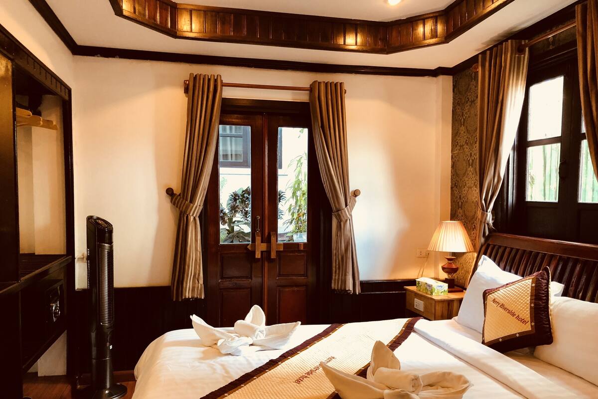 A hotel room with a bed, a bedside table with a lamp, a window with curtains, and a door with a view of the outside.