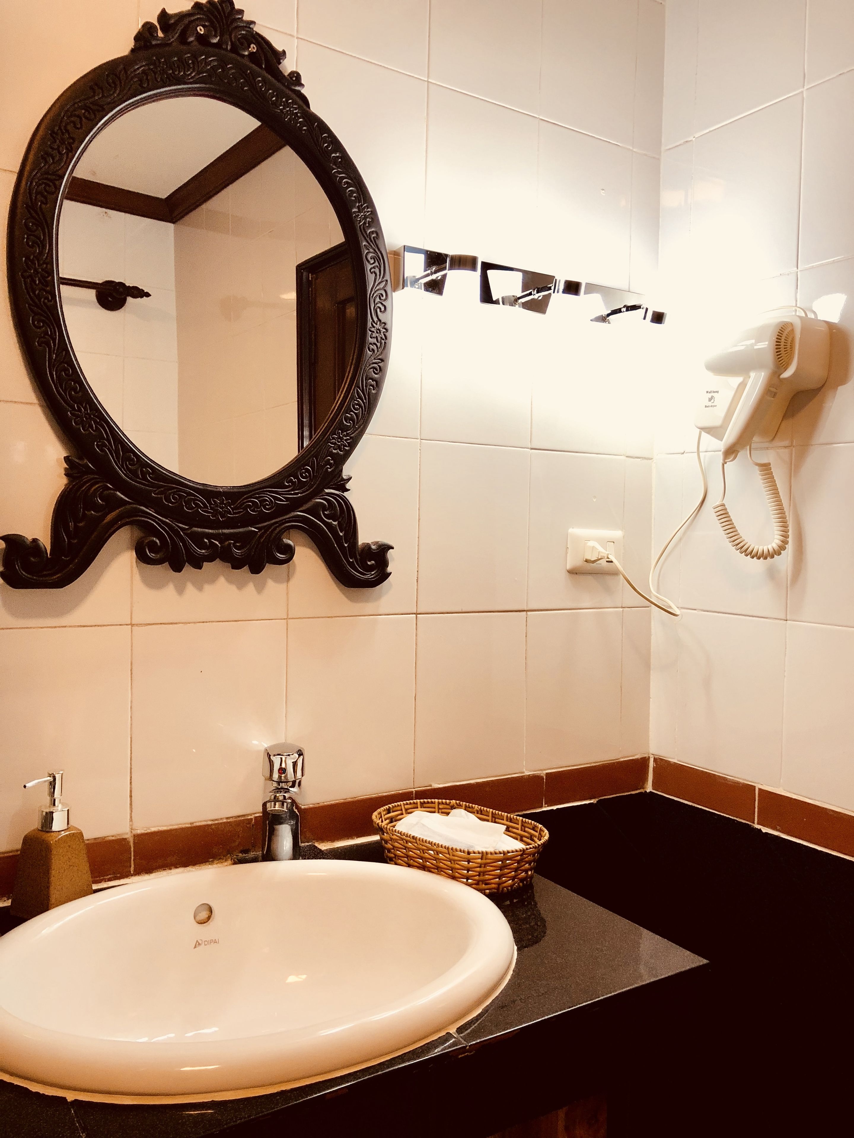 A bathroom with a white sink, a black countertop, a decorative mirror, a hairdryer, and a wicker basket.