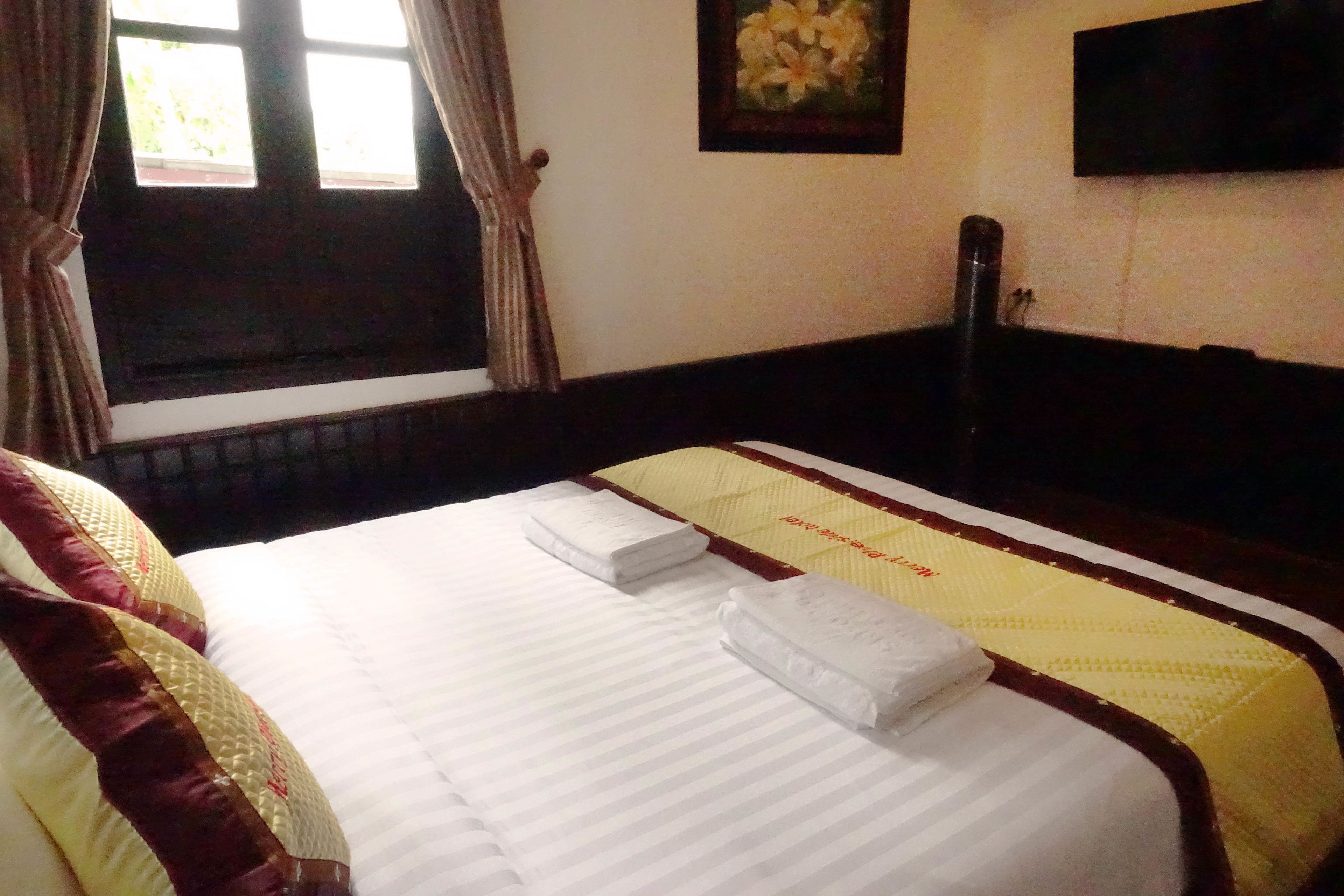 A neatly made bed with white linens and two folded towels, a window with curtains, and a wall-mounted TV.