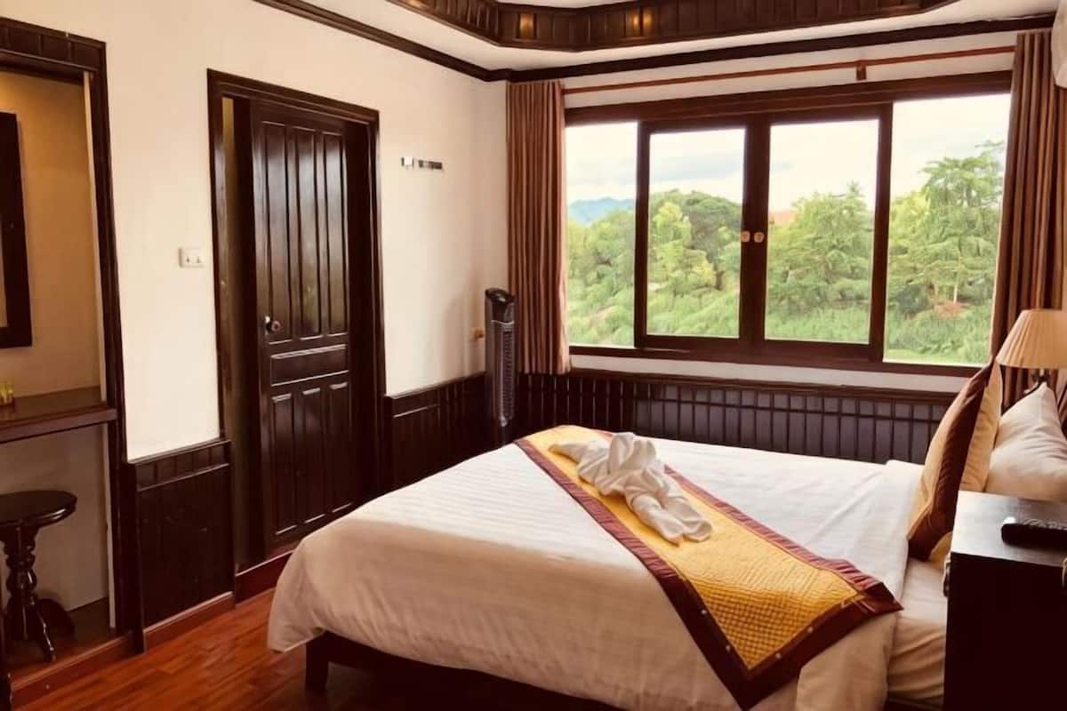 A hotel room with a large bed, a window with a view of greenery, a wooden door, and a bedside table.