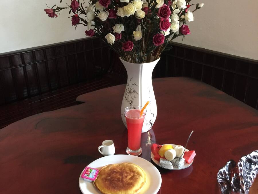 A dining table with a white plate of pancakes, a glass of red juice, a small bowl of fruit, and a vase of flowers.