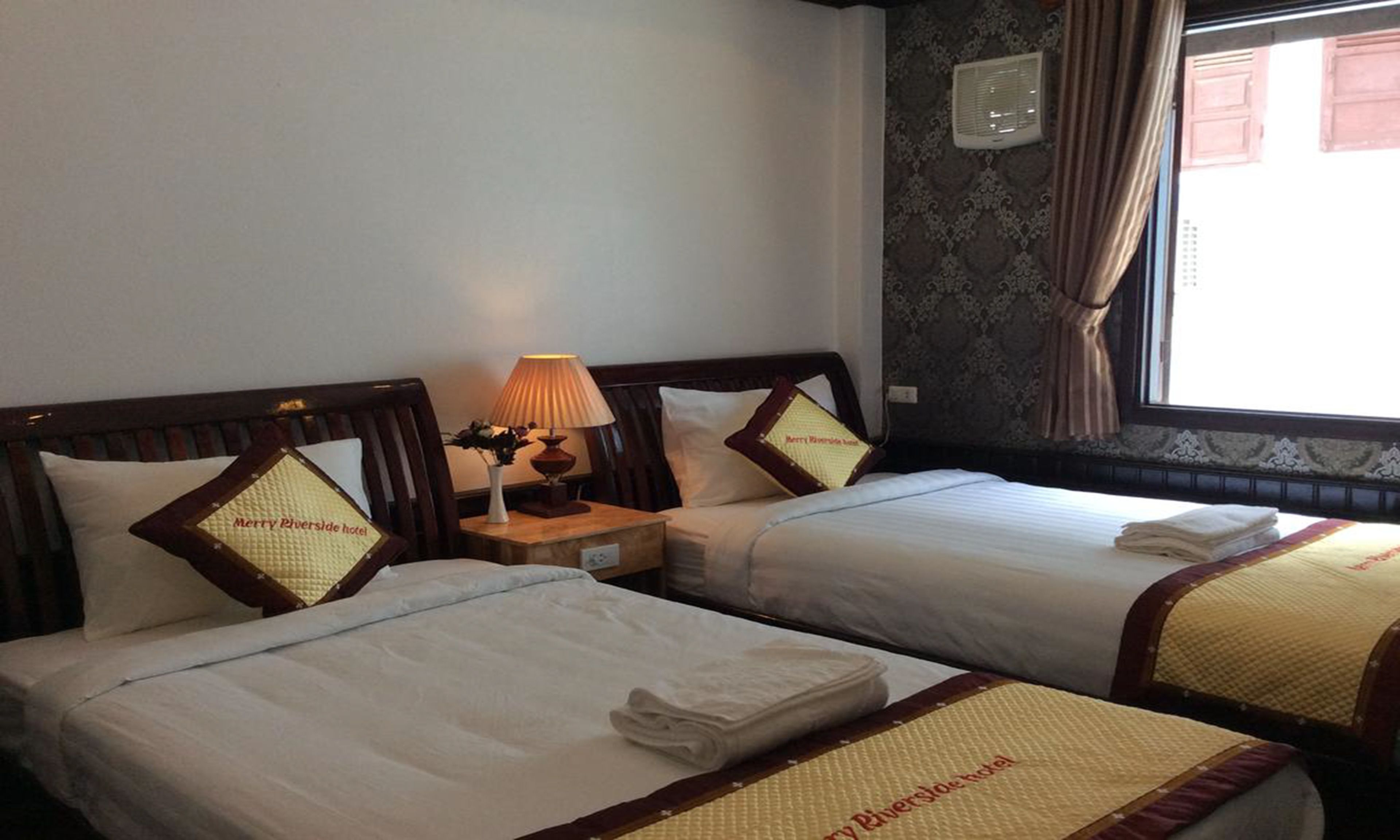 Une chambre d’hôtel avec deux lits, chacun équipé d’un oreiller et d’une couverture, une table de chevet avec une lampe, et une fenêtre avec des rideaux.