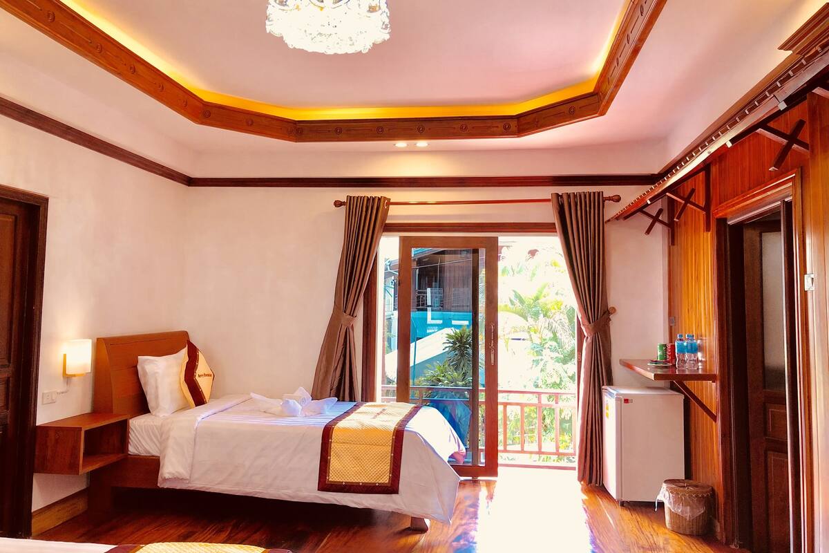 A hotel room with a bed, a desk, a small refrigerator, and a balcony with a view of greenery.