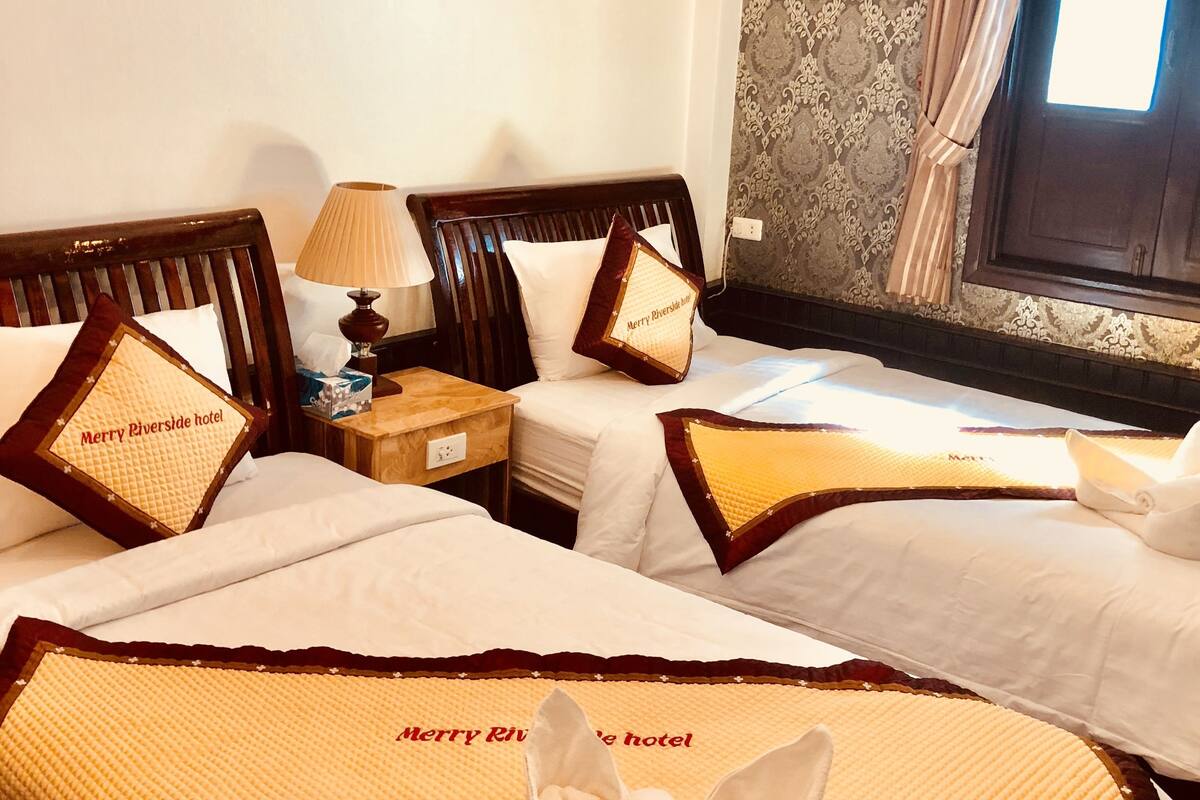 A hotel room with two beds, each with a pillow that reads "Merry Riverside hotel." There is a nightstand with a lamp and a small table.