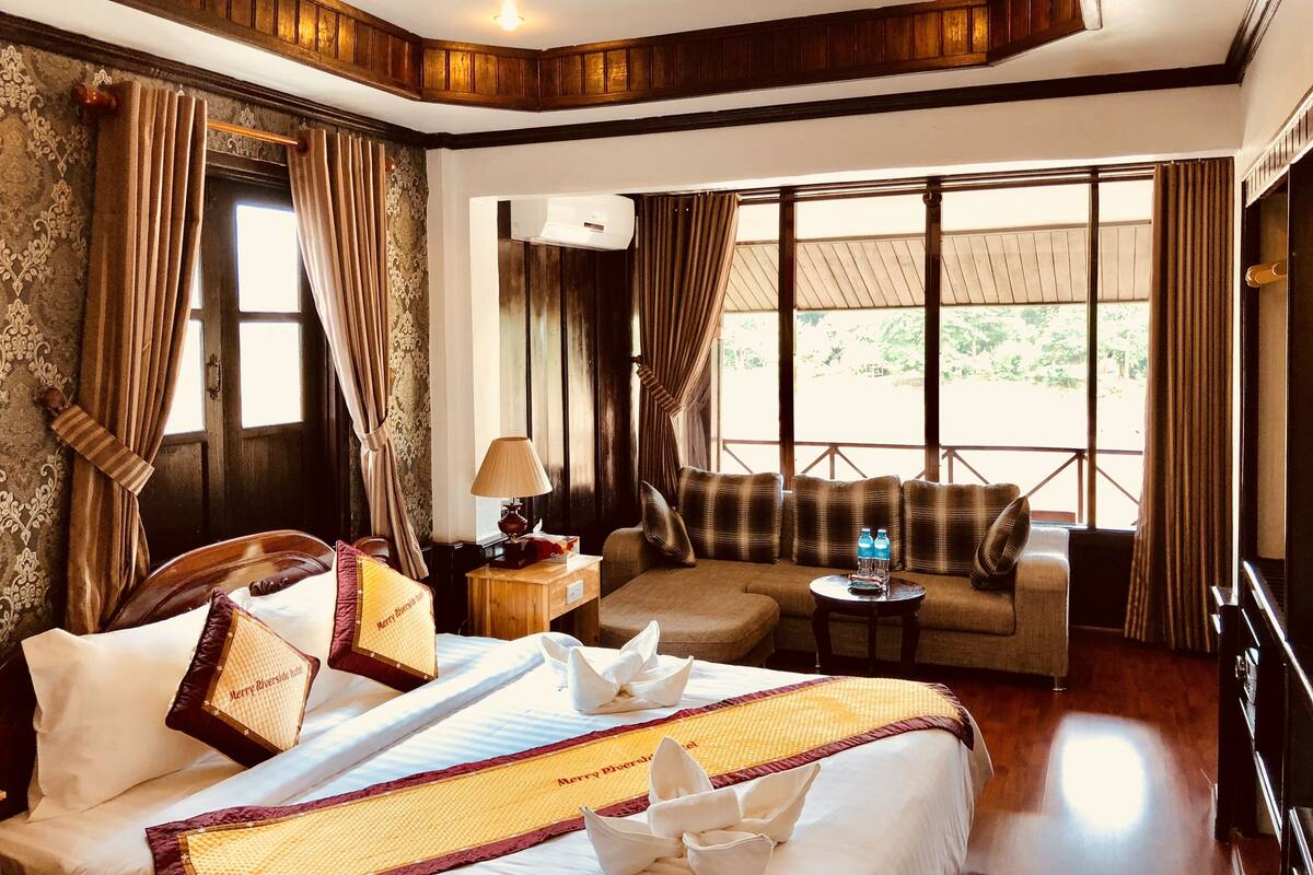A hotel room with a bed, a sofa, a small table with a lamp, and a window with curtains.