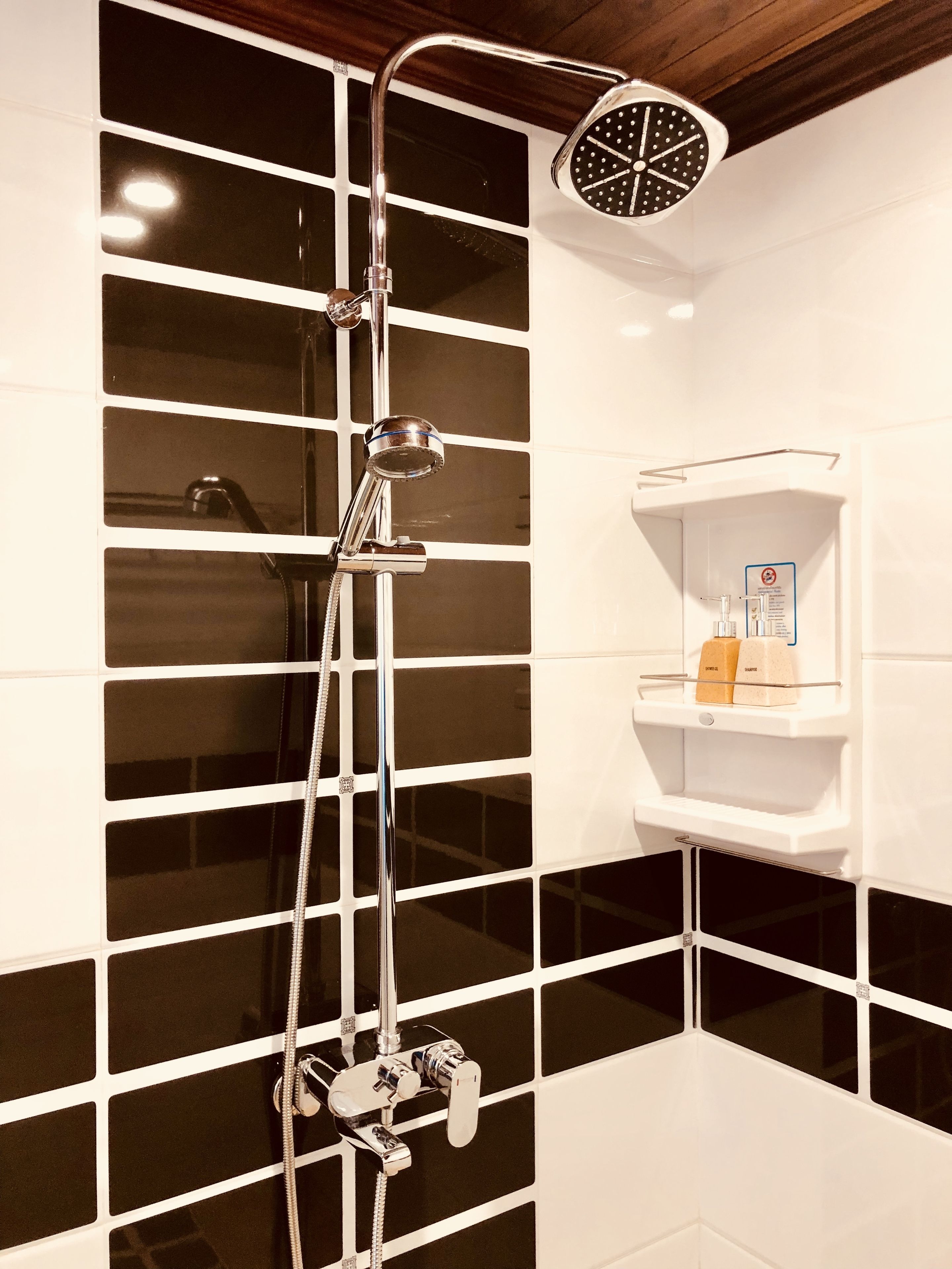 A modern bathroom with a black and white tiled wall, a shower with a handheld showerhead, and a shelf with toiletries.