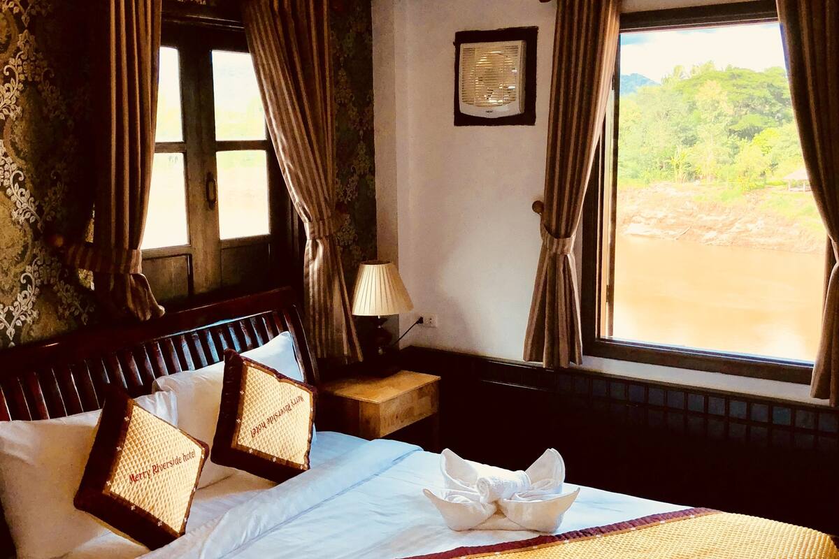 A hotel room with a large bed, a bedside table with a lamp, a window with curtains, and a view of greenery outside.