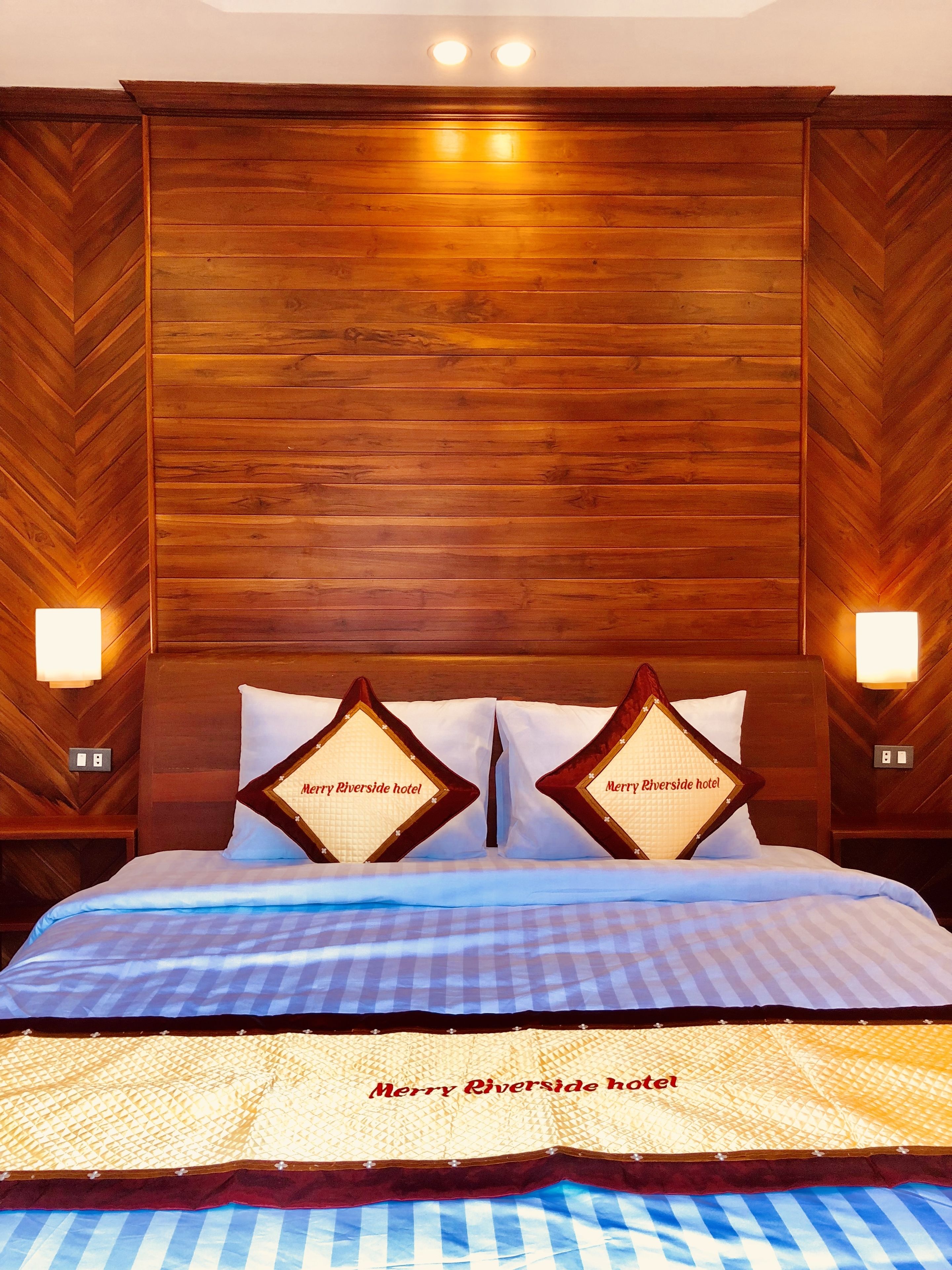 Habitación de hotel con cabecera de madera, dos almohadas con el nombre del hotel, cama con colcha azul y blanca a cuadros, y dos luces fijadas en la pared.