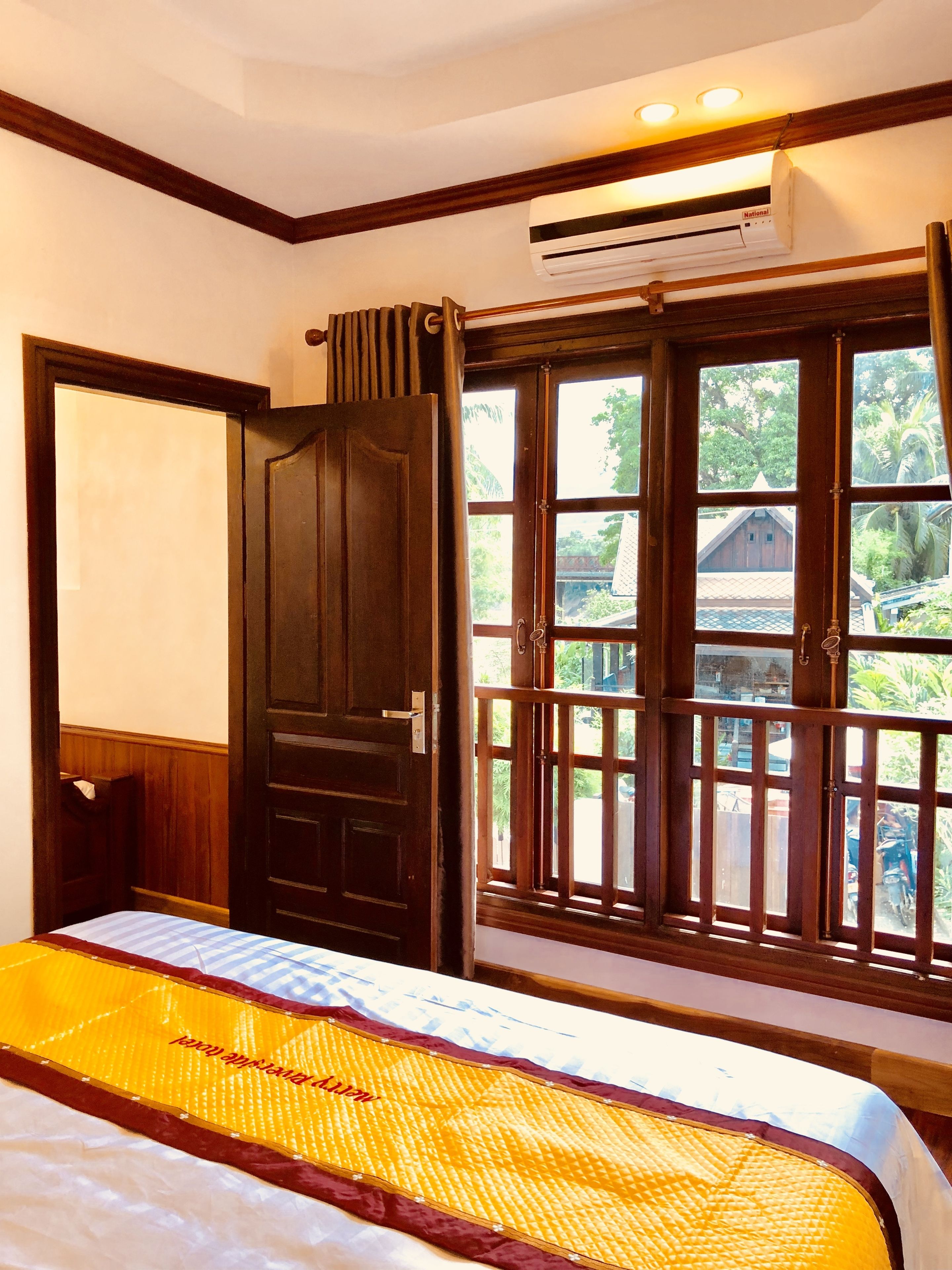 A room with a bed, an air conditioning unit, a window with a view of greenery, and a wooden door.