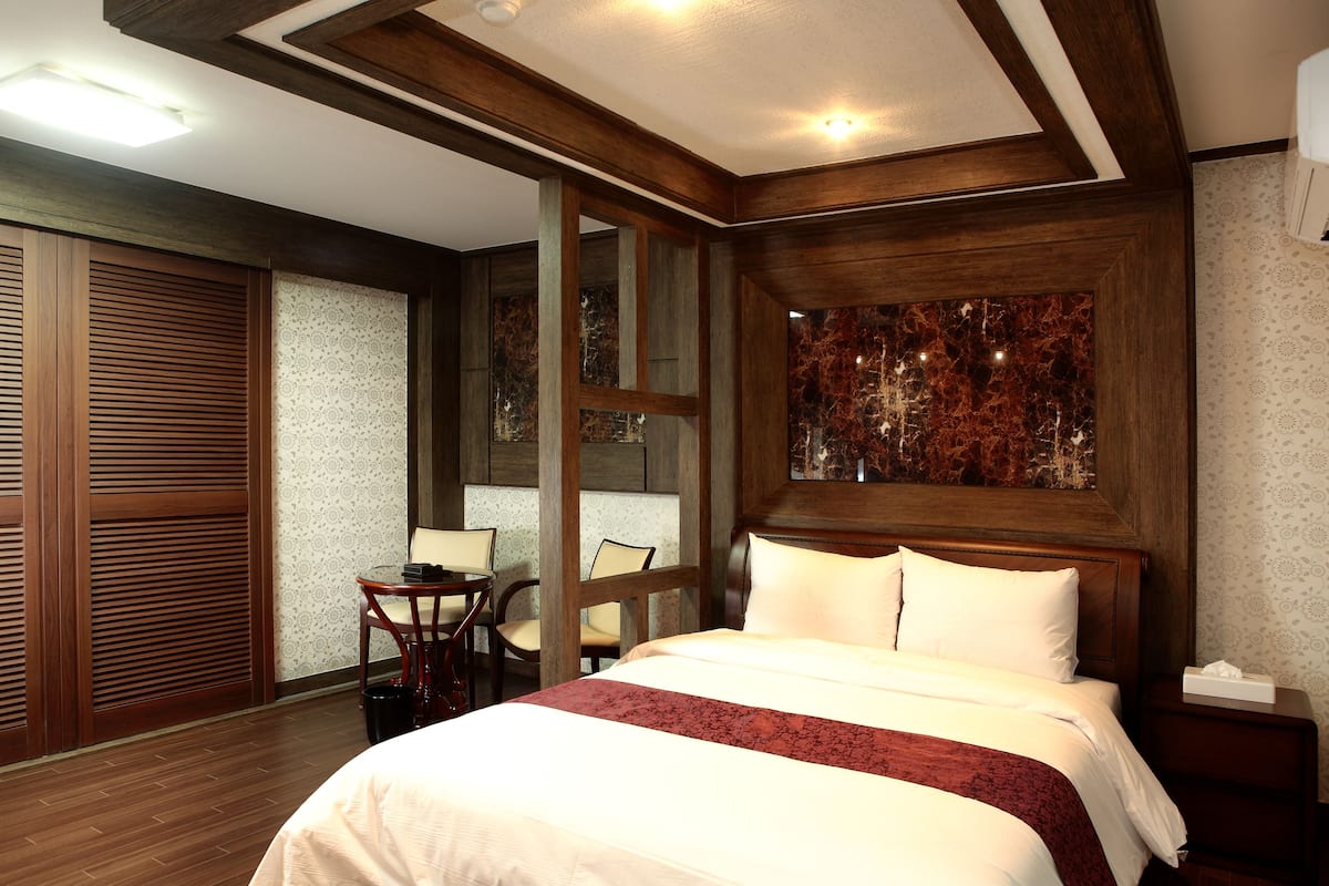 A hotel room with a large bed, wooden headboard, a small table with chairs, and a decorative wall piece.