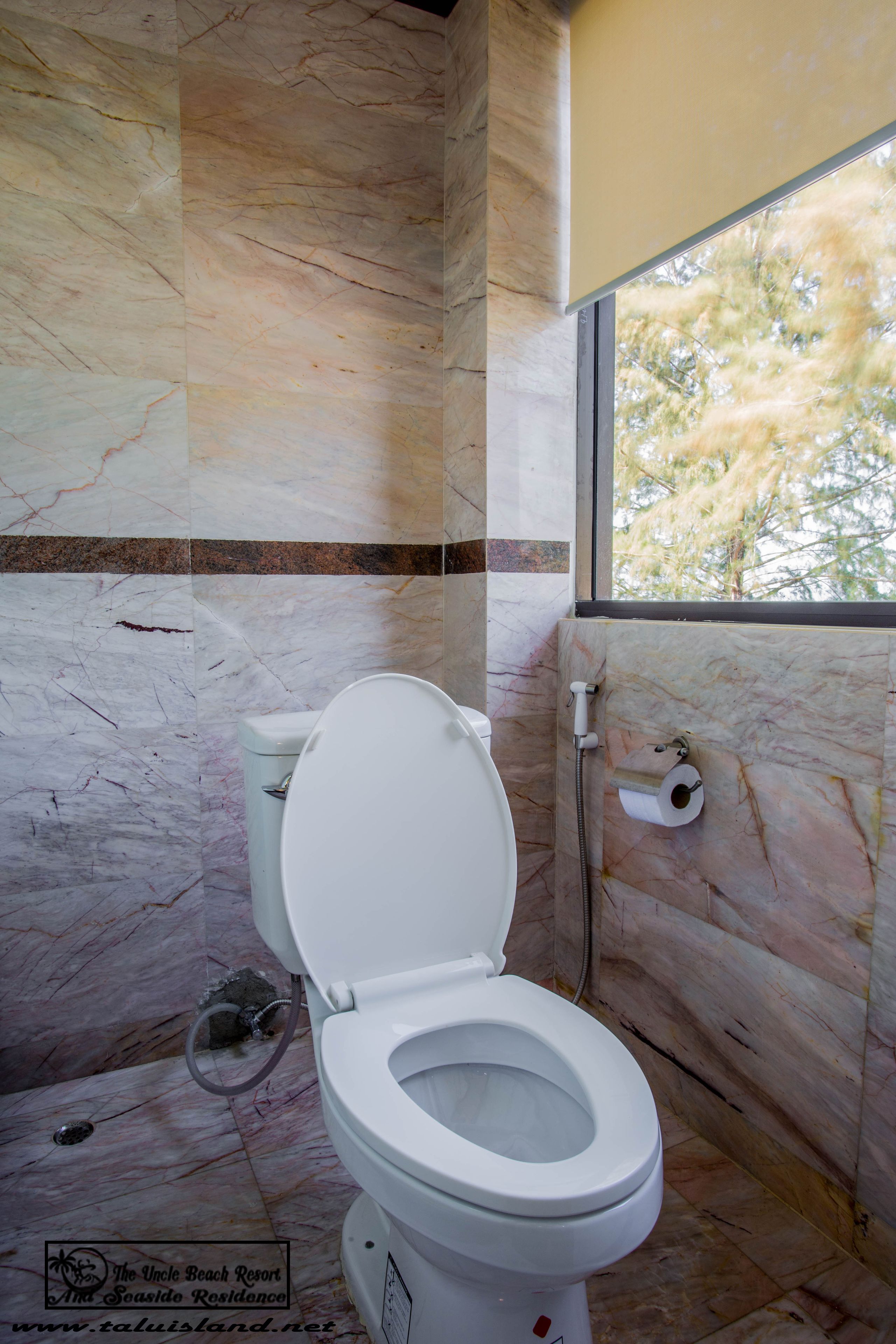 Un baño con pared de mármol, un inodoro y una ventana con vistas a árboles.