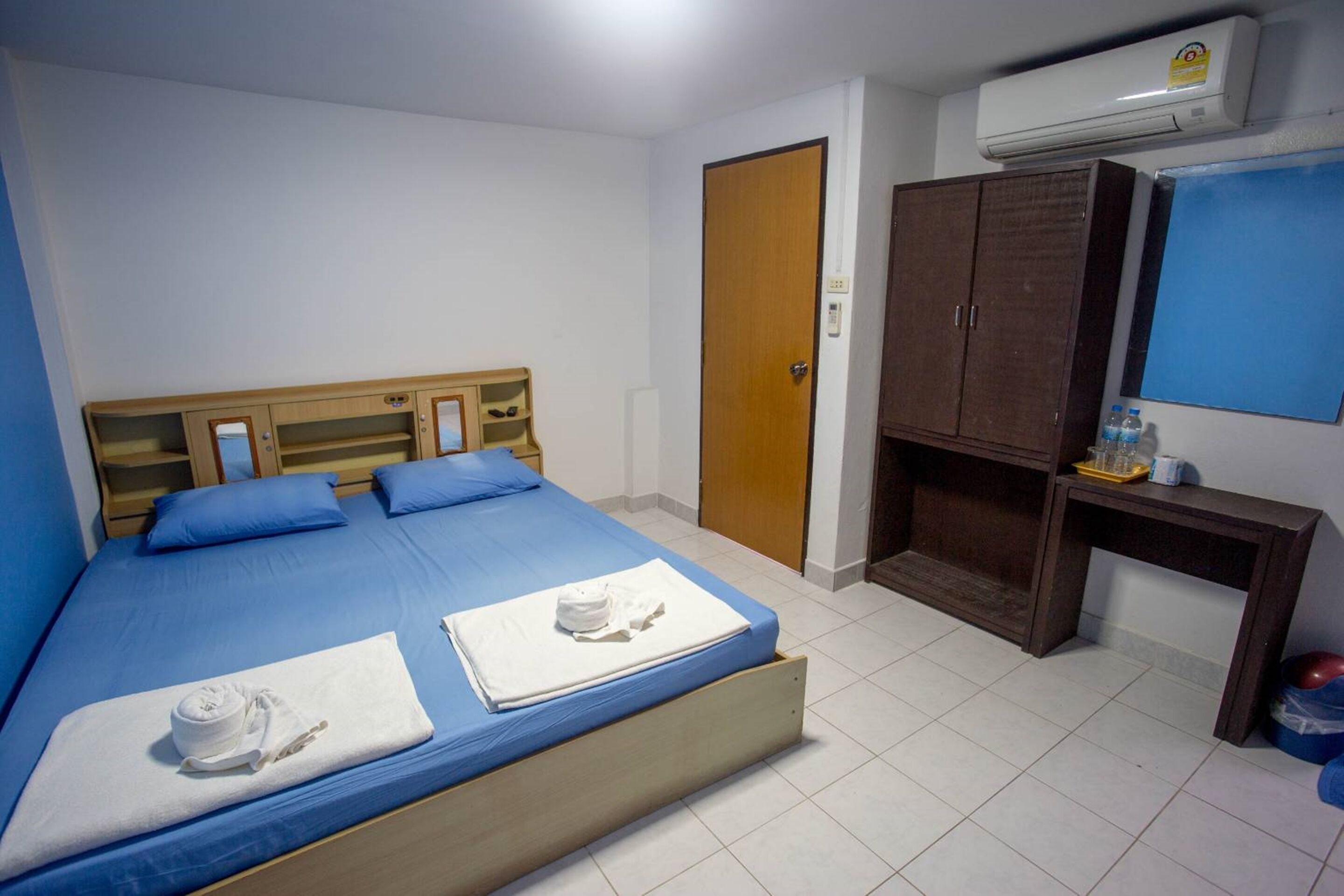 A single bed with blue bedding, a wooden headboard, and two white towels. There is a wooden wardrobe, a small table with a water bottle, and an air conditioning unit on the wall.