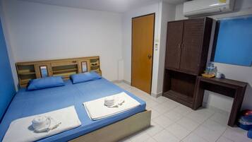 A single bed with blue bedding, a wooden headboard, and two white towels. There is a wooden wardrobe, a small table with a water bottle, and an air conditioning unit on the wall.