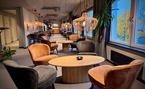 A hotel lobby with round tables, cushioned chairs, hanging wicker lamps, and a large window with a view of trees. - Kramfors Stadshotell (Kramfors)