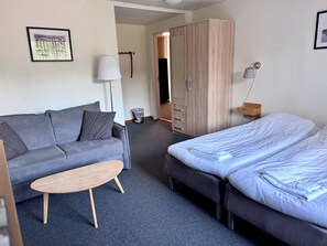 Habitación de hotel con un sofá gris, una mesa de centro de madera, una cama con ropa blanca, un armario y un cuadro en la pared.