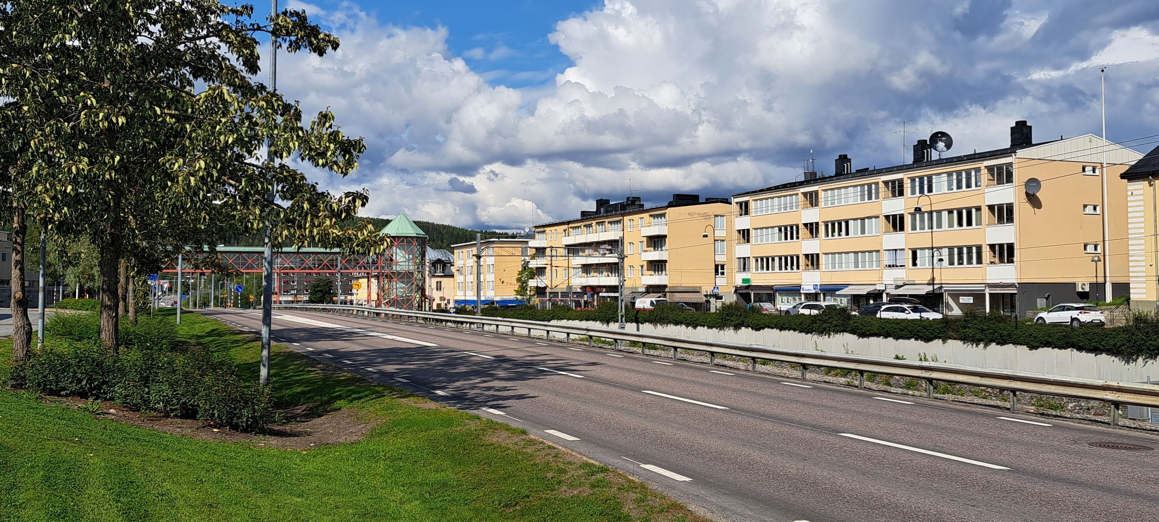 Un immeuble résidentiel à plusieurs étages, doté de balcons, d’un espace de stationnement et bordé d’une route protégée par des garde-fous.