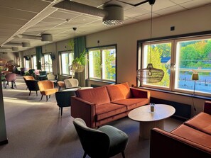 A modern lounge area with a sofa, chairs, a round table, and large windows with a view of greenery. - Kramfors Stadshotell (Kramfors)