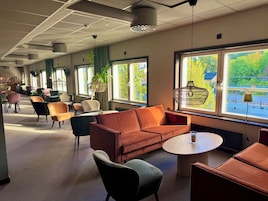 Espaço de estar moderno com sofá, cadeiras, mesa redonda e amplas janelas com vista para o verde.
