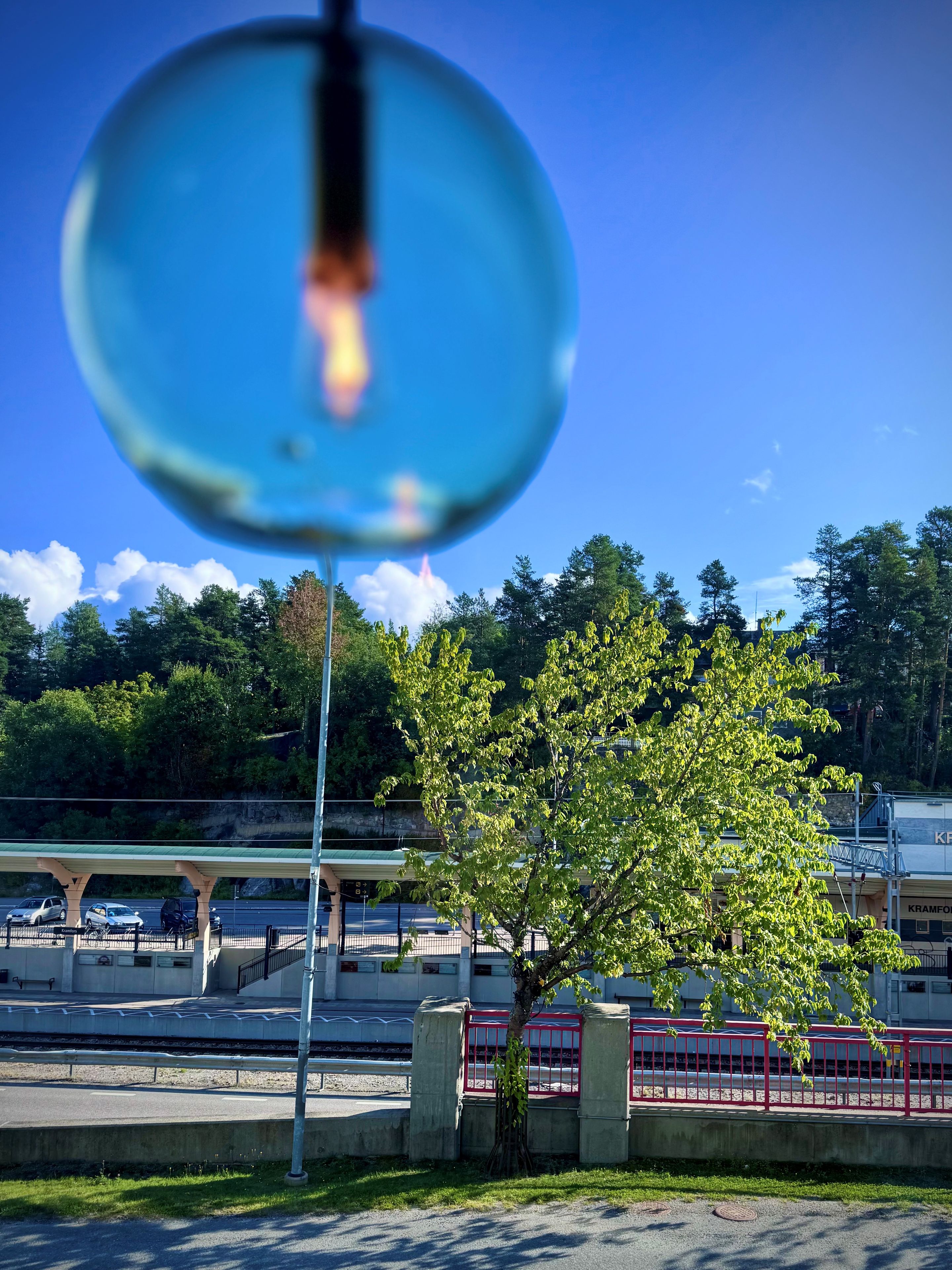 A clear view of a train station platform with a tree in the foreground and a building with 'KRAMFO' signage in the background.