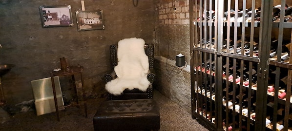 Una habitación pequeña y tenuemente iluminada con un sillón antiguo cubierto con una manta de piel blanca, una botella de metal llena de botellas de vino y una mesita con una vela.