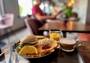 Bandeja de desayuno con waffles, un sándwich, un vaso de jugo de naranja y una taza de café.
