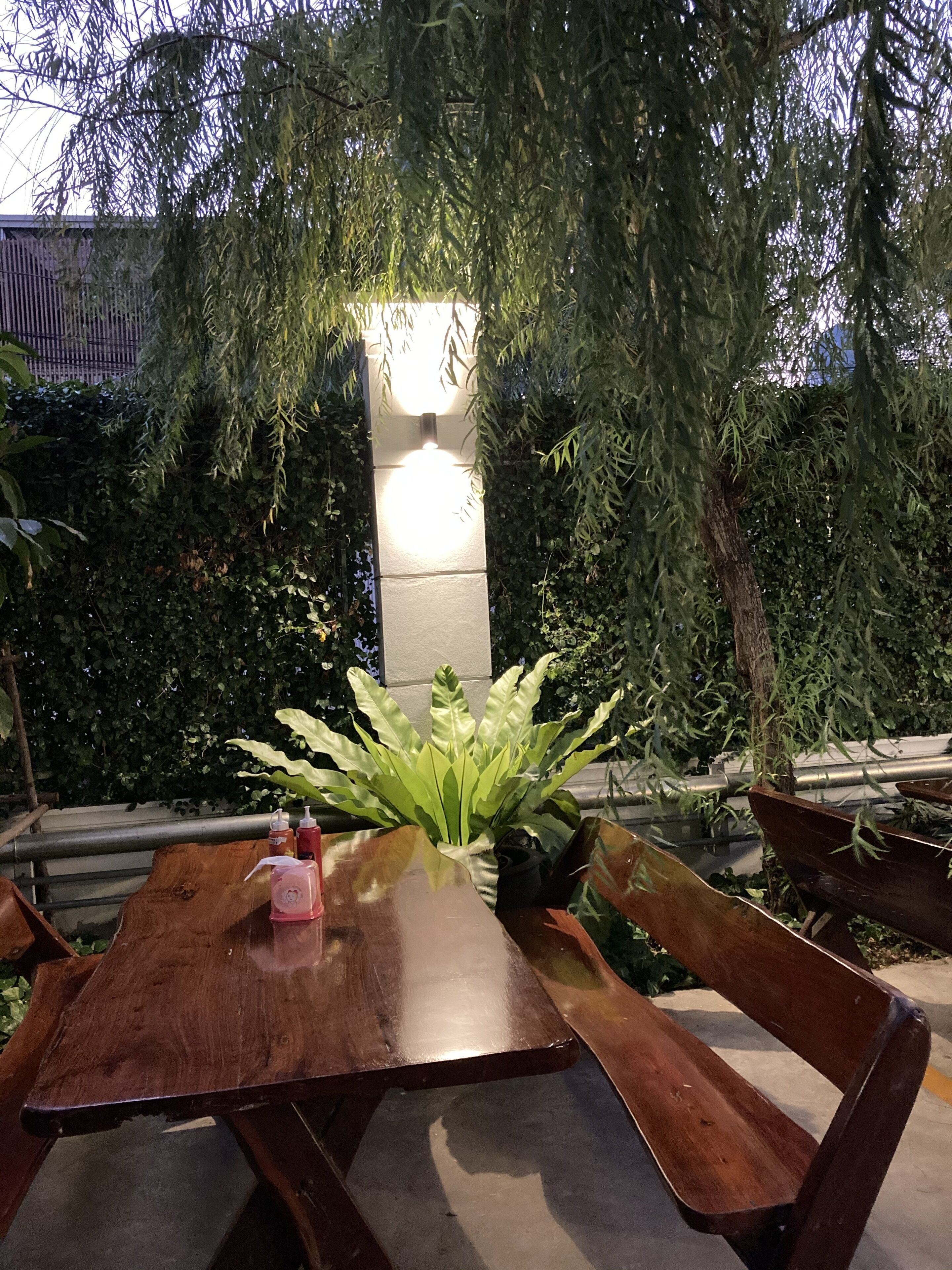 Une table en bois installée à l’extérieur, avec des bancs, entourée d’une végétation luxuriante et agrémentée d’un pilier en pierre surmonté d’un luminaire.
