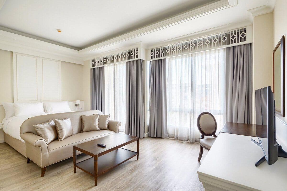 A modern hotel room with a beige sofa, a wooden coffee table, a desk with a computer, and large windows with grey curtains.