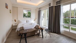 A hotel room with a large bed, a sofa, a small table, and a chair. There are large windows with curtains, a ceiling light, and a wall-mounted TV. - Siam Tharadol (Bangkok)