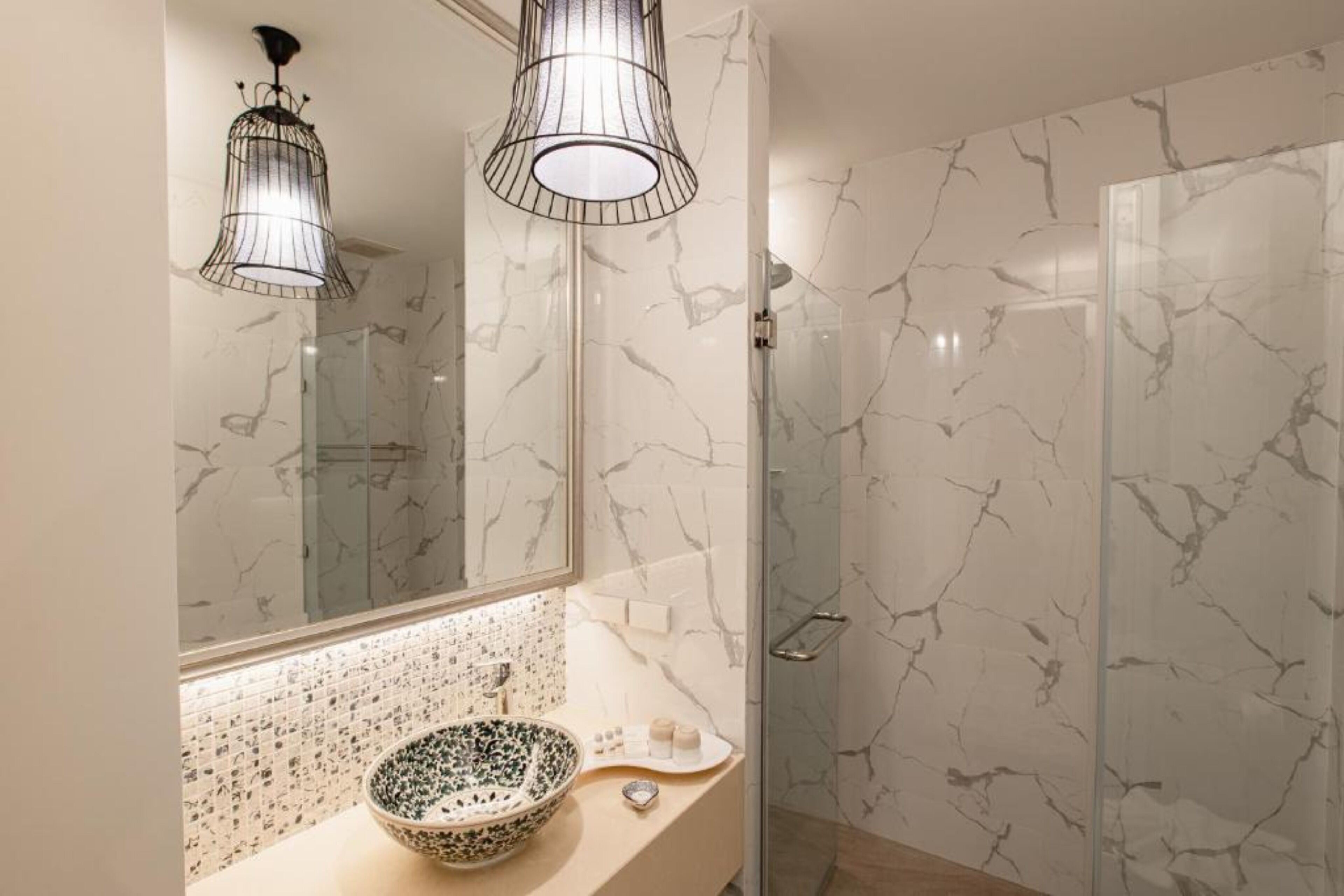 A bathroom with a marble backsplash, a bowl sink, a large mirror, and a glass shower enclosure.
