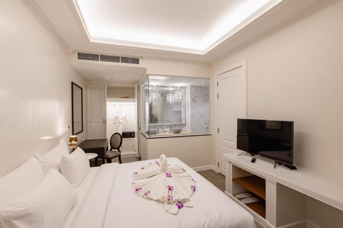 A hotel room with a bed, a TV, a desk, and a bathroom visible through a glass door.