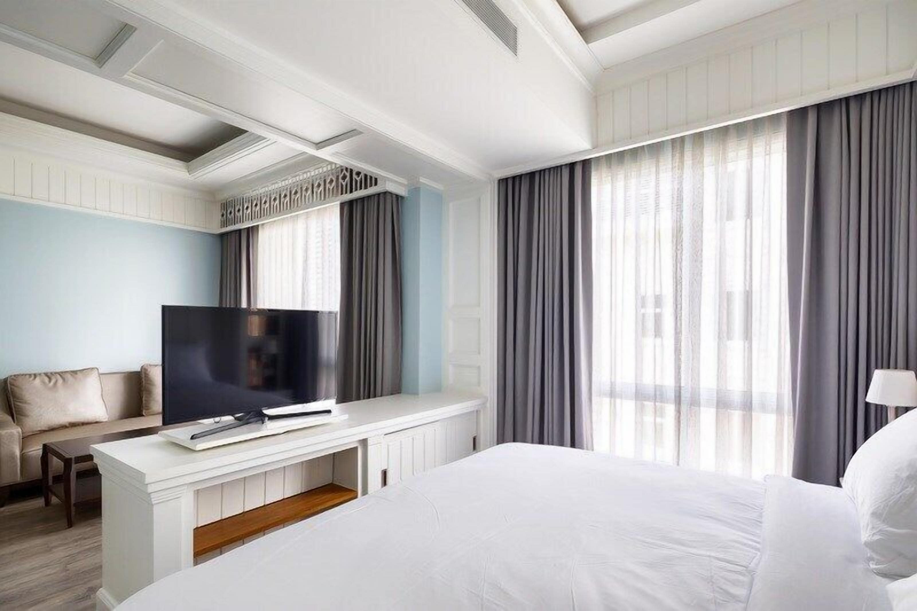 Ein Hotelzimmer mit Bett, Fernseher auf einem weißen Konsolentisch, einer Couch und großen Fenstern mit Vorhängen.