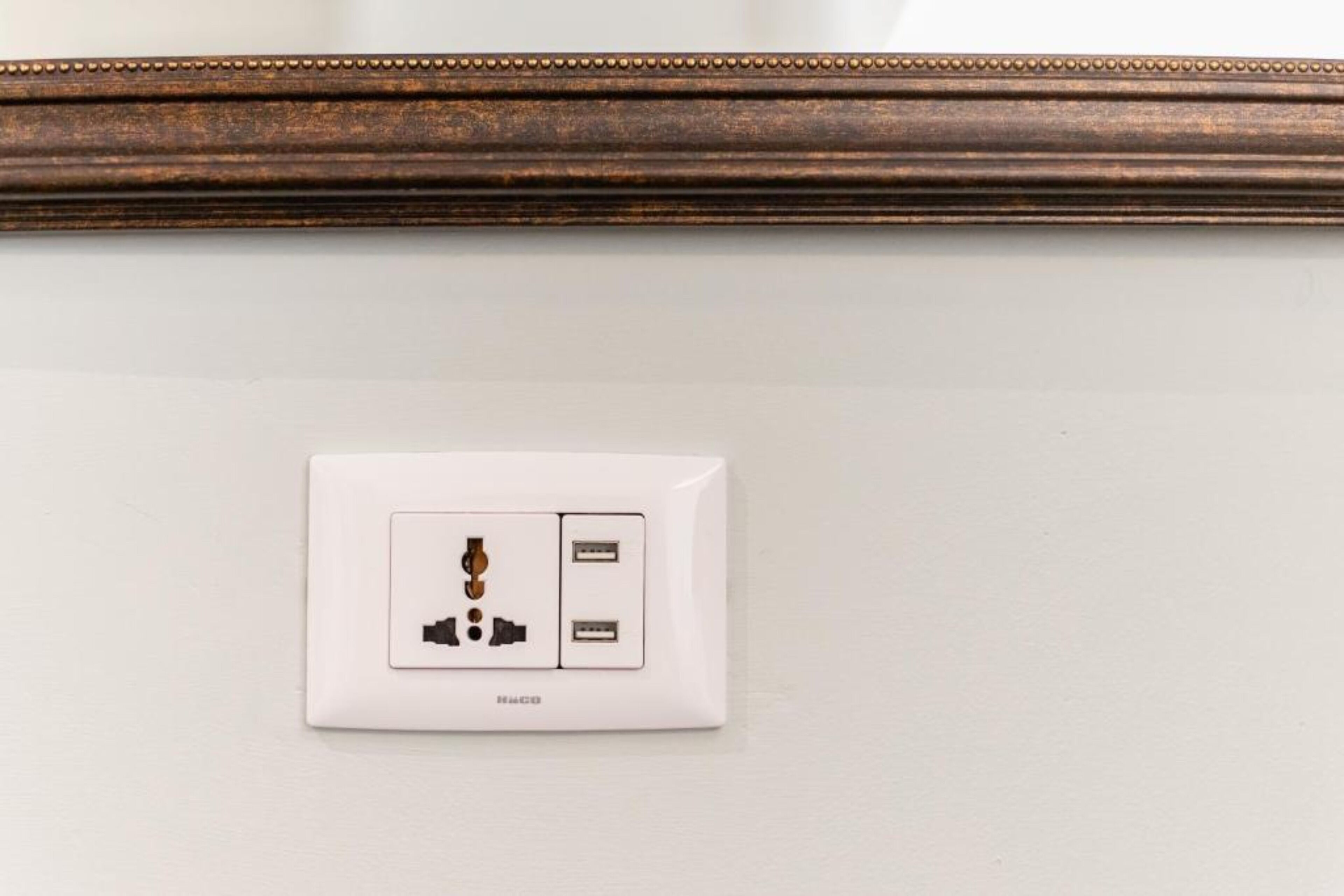 Un enchufe blanco con un puerto USB y un enchufe estándar, instalado en una pared blanca sobre un listón de madera.