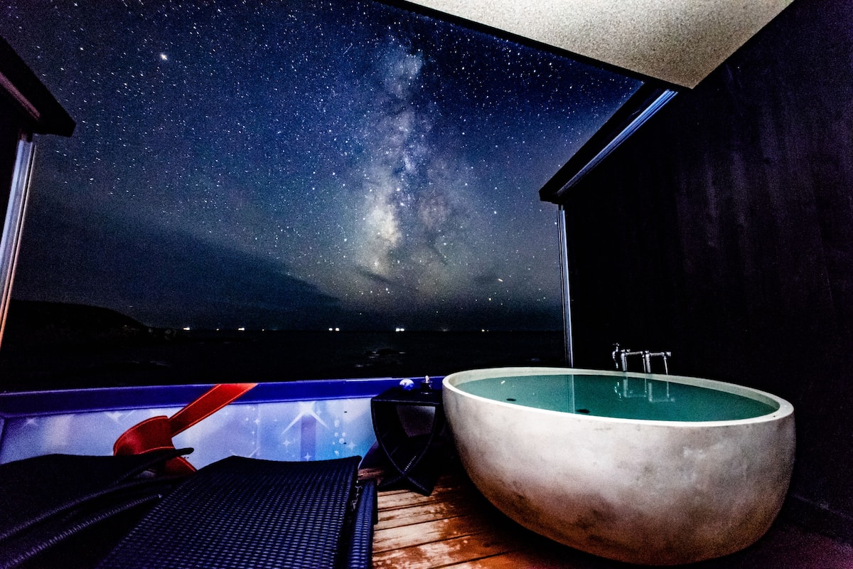 A room with a large window revealing a starry night sky, a jacuzzi, and a wooden floor.