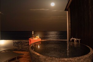 Un jacuzzi con vista al océano iluminado por la luna, una botella y copas sobre una pequeña mesa.