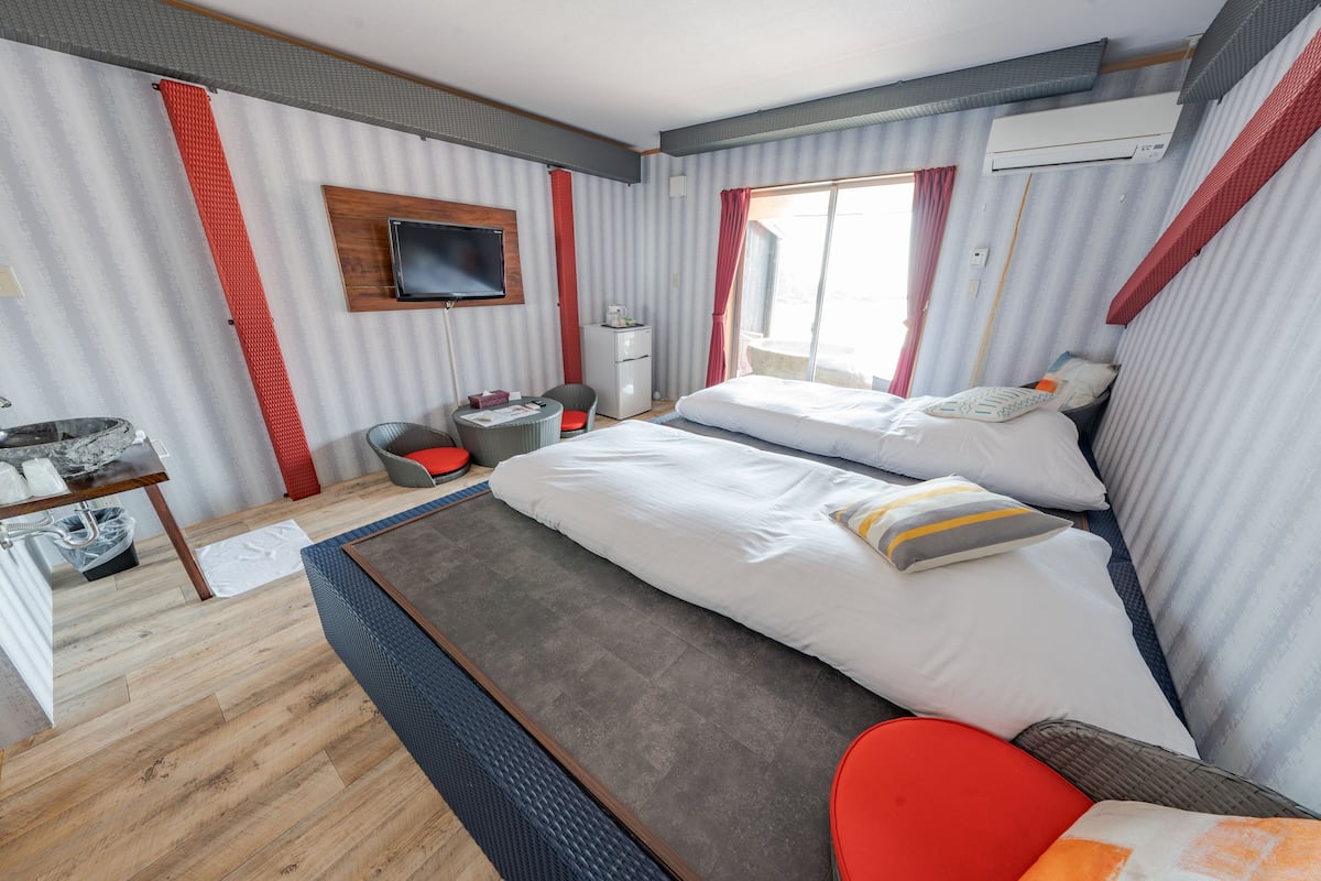 A hotel room with two beds, a TV, a desk with a chair, a small table, and a window with curtains.