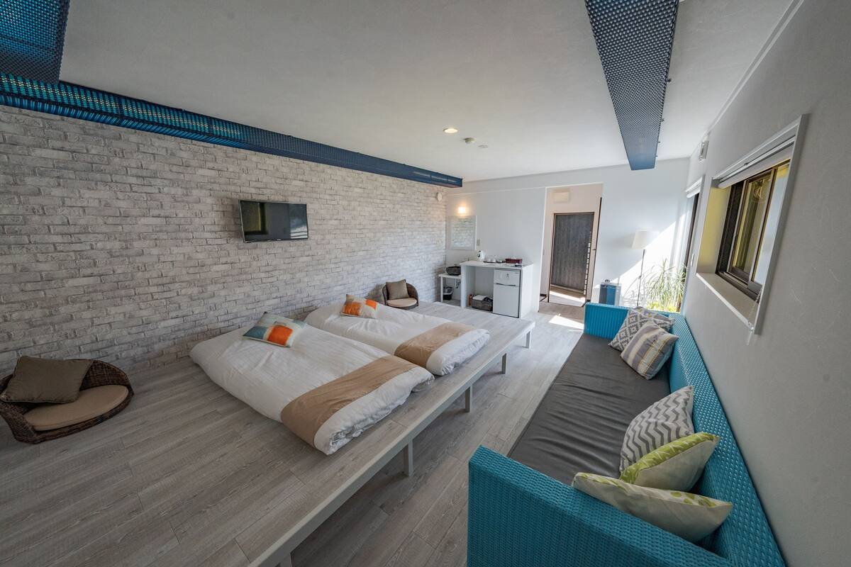 A modern hotel room with a brick wall, two beds, a sofa, a TV, and a small kitchenette.