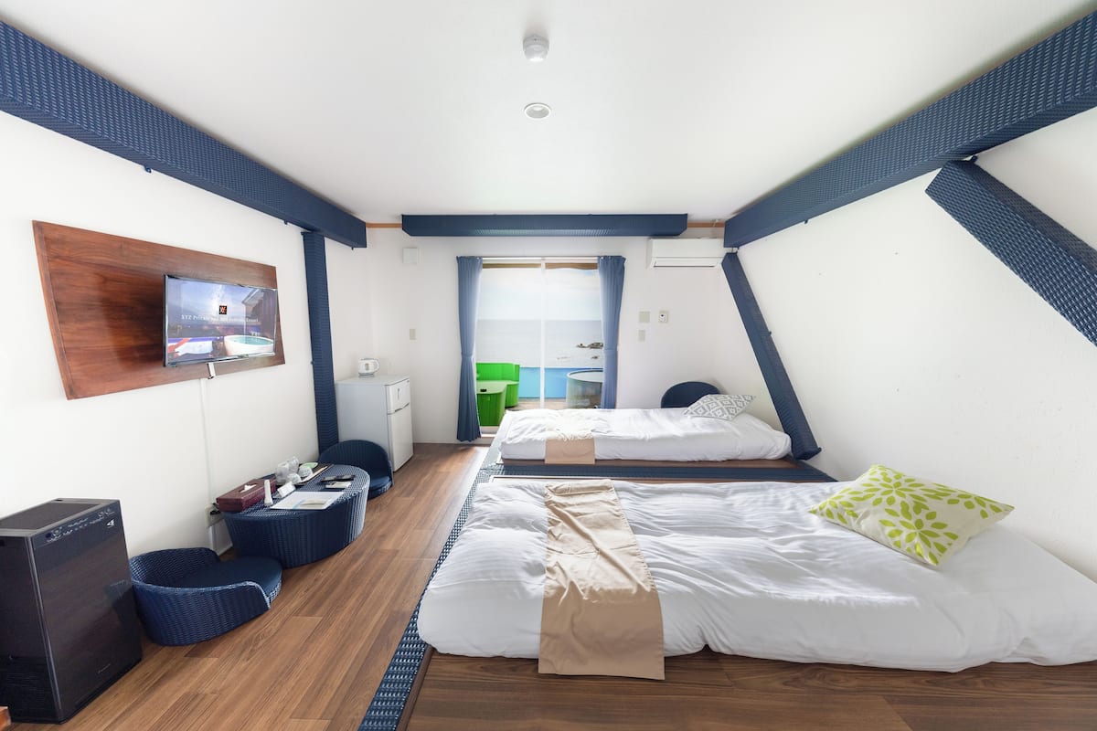 A room with two beds, a TV on the wall, a desk with a computer, and a window with curtains.