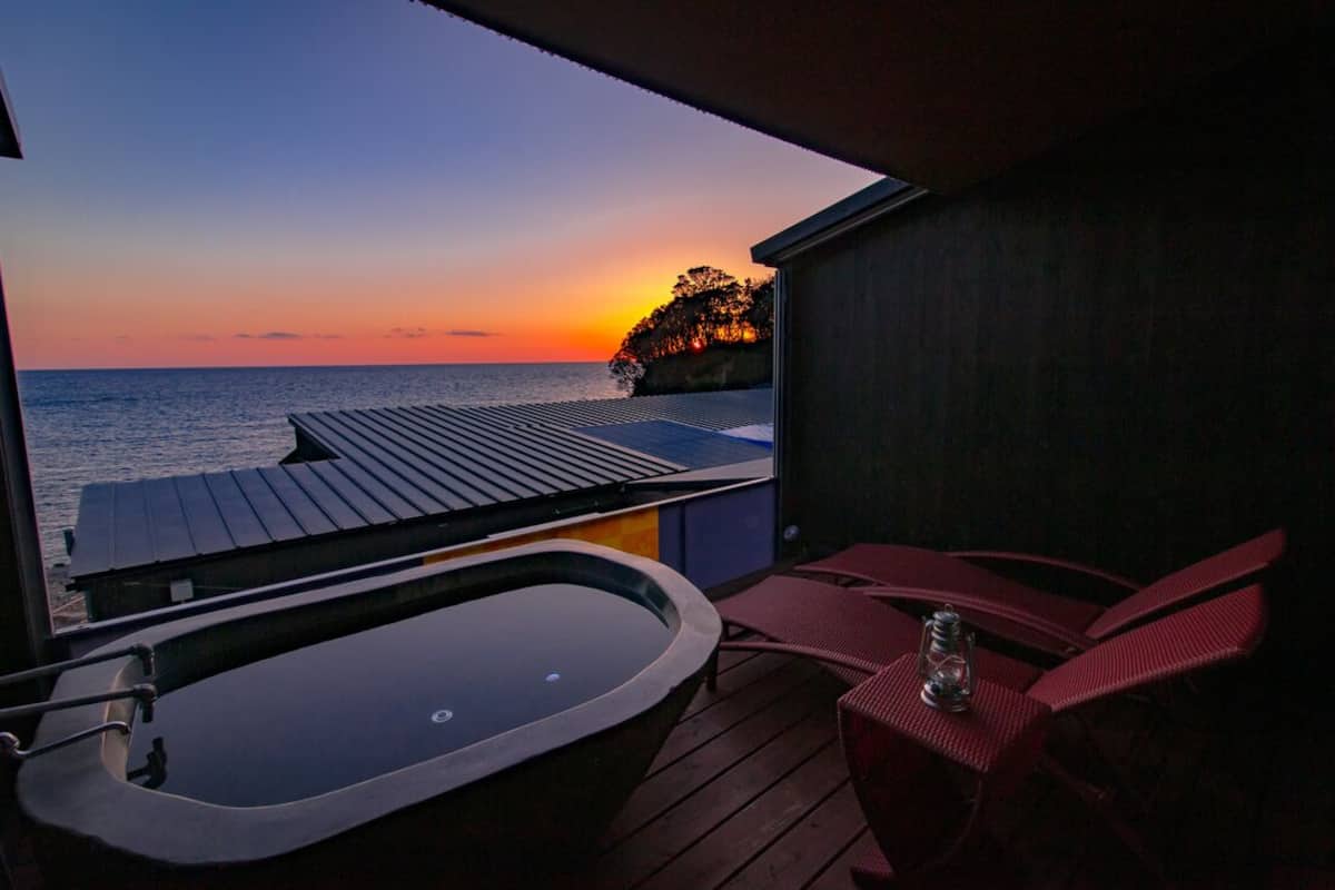 A balcony with a view of the sunset over the sea, a hot tub, and red lounge chairs.