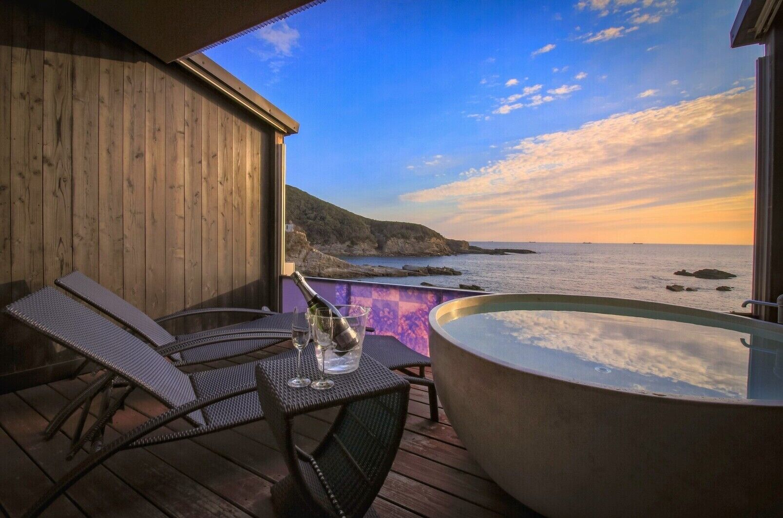 Een houten terras met een jacuzzi, lounge stoelen en uitzicht op zee bij zonsondergang.