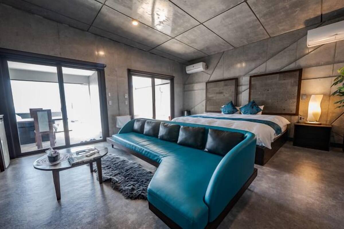 A modern hotel room with a large bed, a blue sofa, a small table, and a large window.
