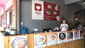 Un restaurante con un mostrador de madera que exhibe varios platos, un menú en la pared con ilustraciones de comida y una persona dando un pulgar arriba.