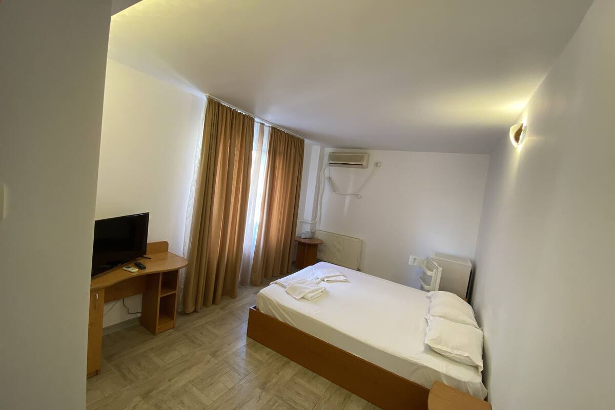 Quarto de hotel com cama, escrivaninha com televisão, janela com cortinas, radiador e ar condicionado.