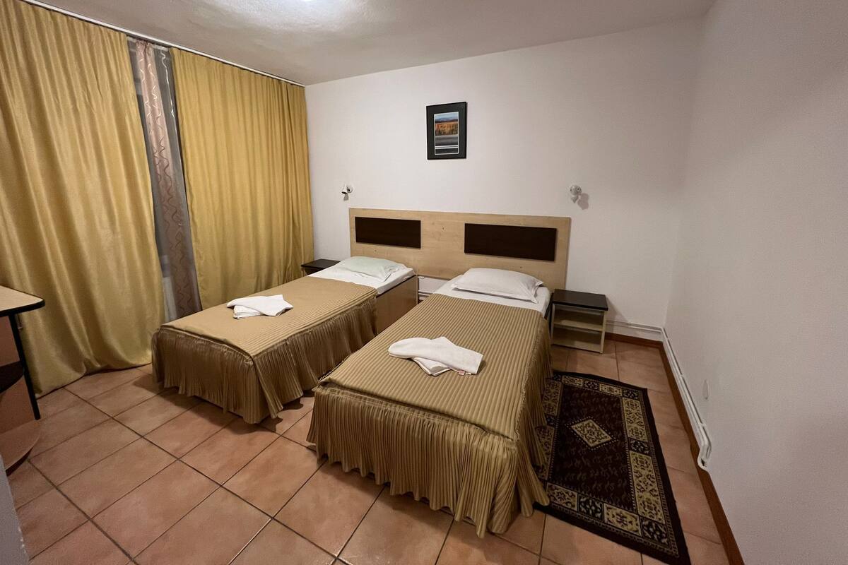 Quarto de hotel com duas camas de solteiro, uma mesa pequena, uma cadeira, uma janela com cortinas e um quadro na parede.