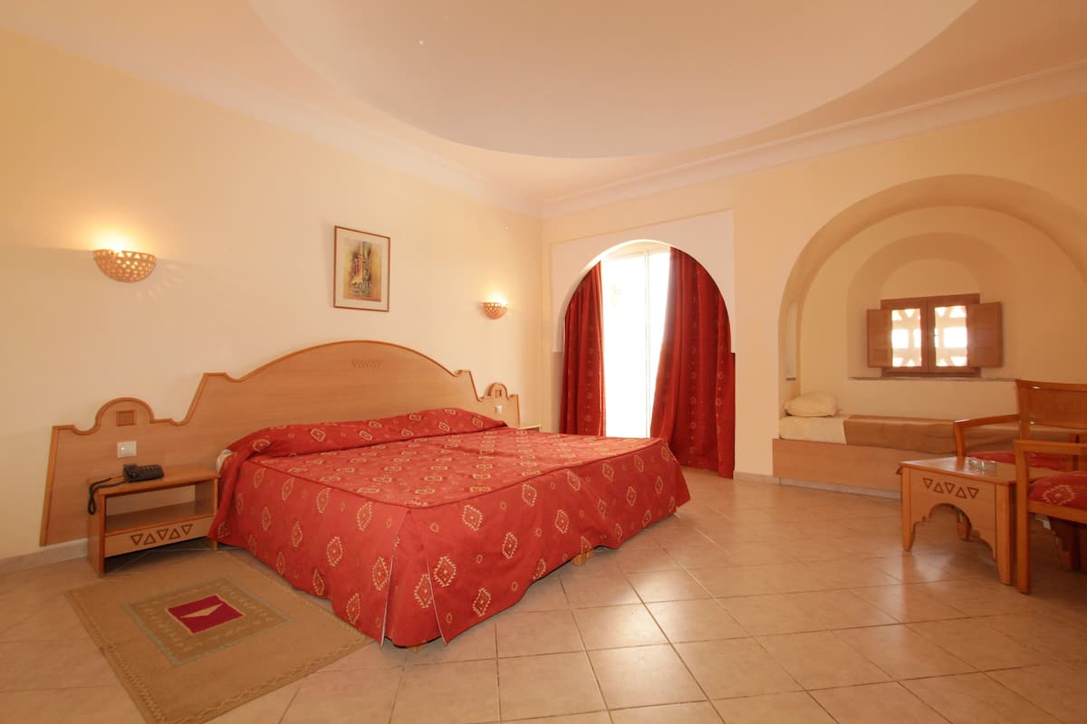 Habitación de hotel con cama de madera, mesita de noche, colcha roja, sofá y mesa de madera.