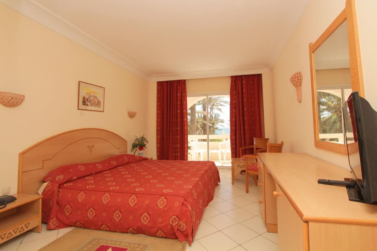 Habitación de hotel con cama, escritorio, silla, televisor y balcón con vista a palmeras.
