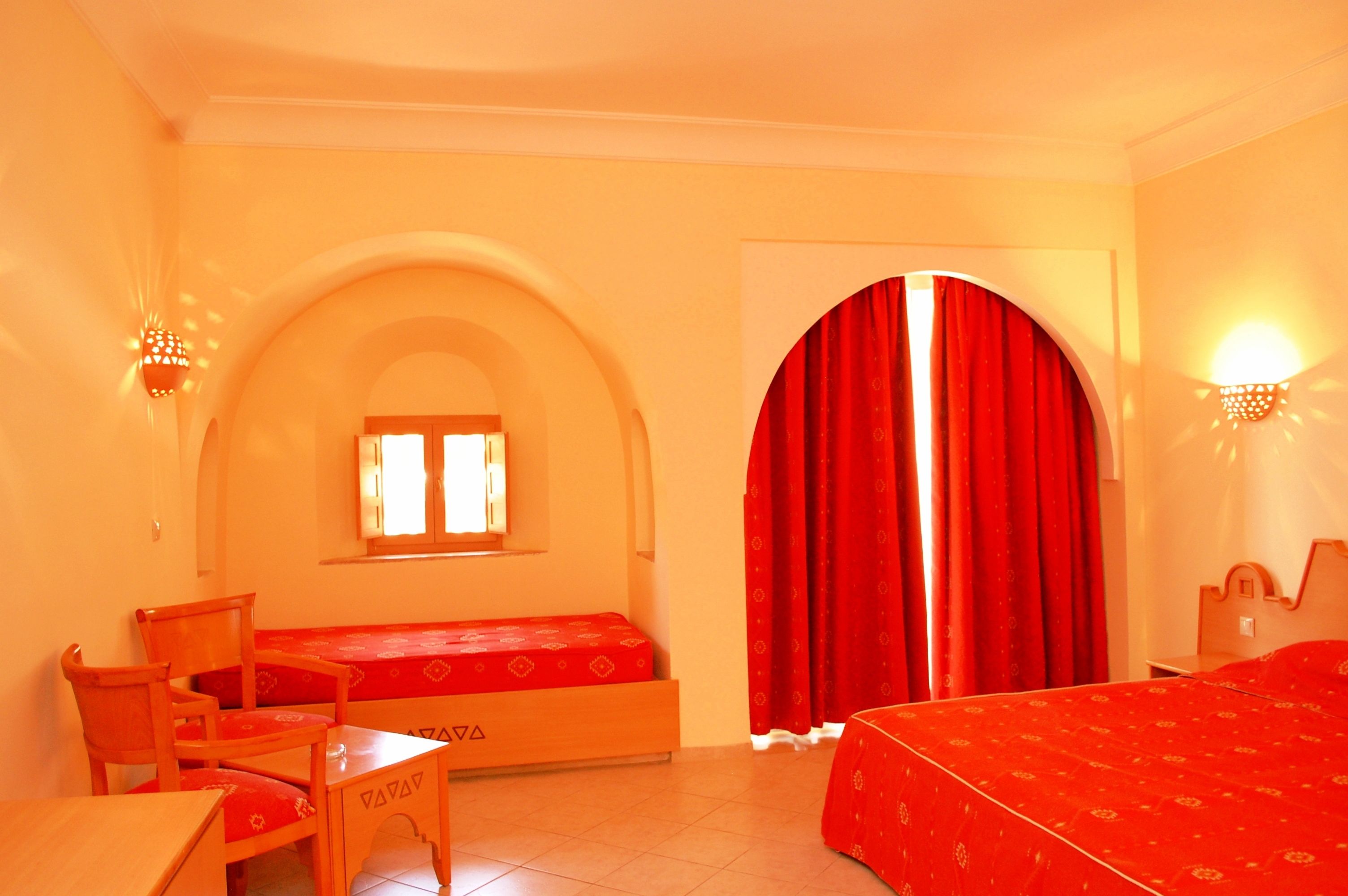 A room with a bed, a desk, and a chair. There is a window with curtains, a wall-mounted light, and a door with a curtain.