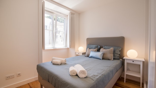 A neatly made bed with a grey bedspread and two rolled white towels, a bedside table with a lamp, and a window with a view of buildings. - Alfama Premium by Homing (Lisbon)
